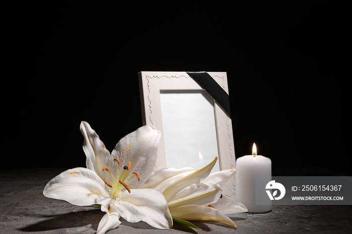 Blank funeral frame, burning candle and flowers on table against dark background