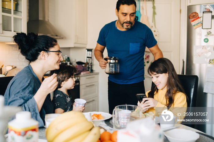 Family looking at smiling girl using mobile phone during breakfast at home