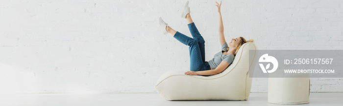 panoramic shot of cheerful blonde girl lying on  bean bag chair and gesturing near brick wall