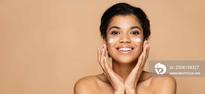 joyful african american woman with face cream on cheeks isolated on beige, banner