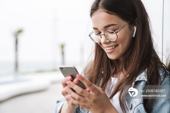 Smiling young pretty woman student wearing eyeglasses walking outdoors listening music with earphone