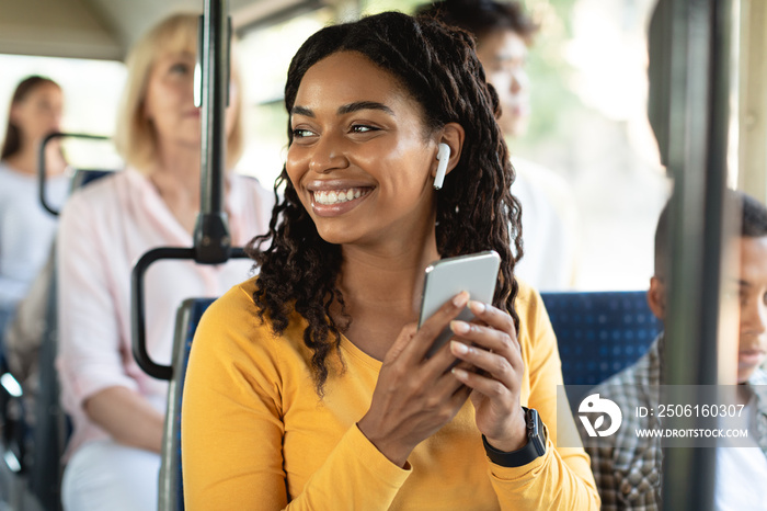 Cheerful black lady using smartphone wearing headphones in bus