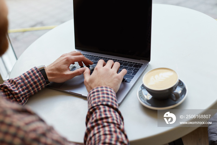 Mens hands are working on the keyboard of the laptop, nearly a gray cup of coffee on a white table.