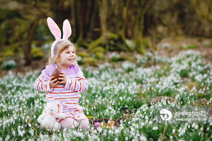 Little girl with Easter bunny ears making egg hunt in spring forest on sunny day, outdoors. Cute hap