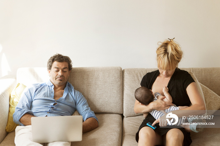 Mother breast feeding her daughter beside father using laptop