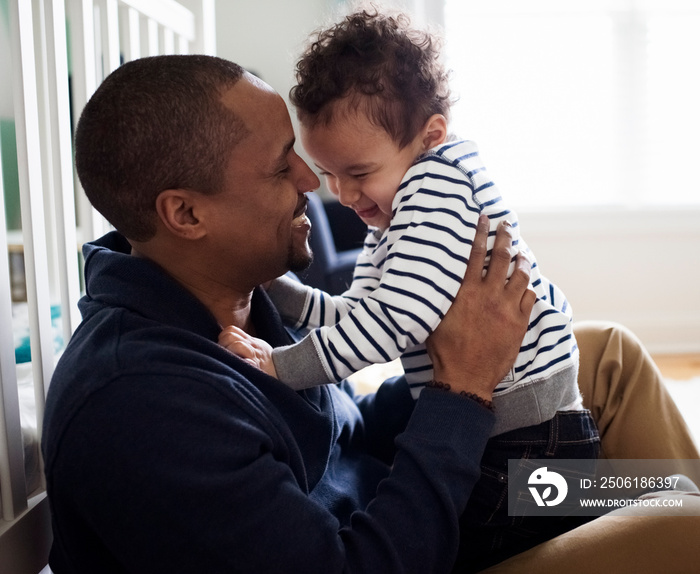 Side view of happy father holding cute son while sitting by crib against window at home