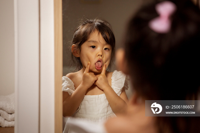 Rear view of cute girl sticking out tongue while looking in mirror at home