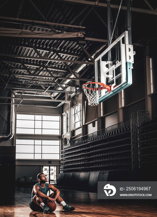 Portrait of Black basketball player sits on a floor under  a hoop in a basketball hall.