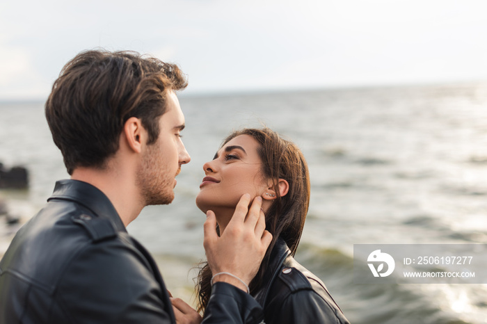 Young man touching cheek of brunette girlfriend in leather jacket on beach