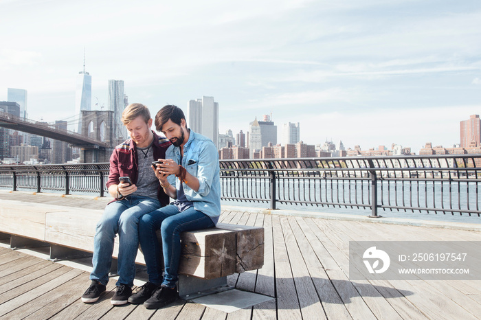 Male couple sitting on riverside by Brooklyn Bridge reading smartphones, New York, USA