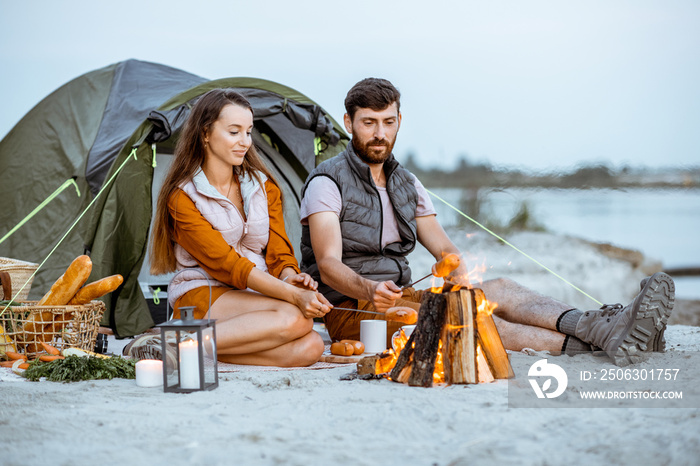 Young and cheerful couple sitting at the fireplace, cooking sausages, having a picnic at the campsit