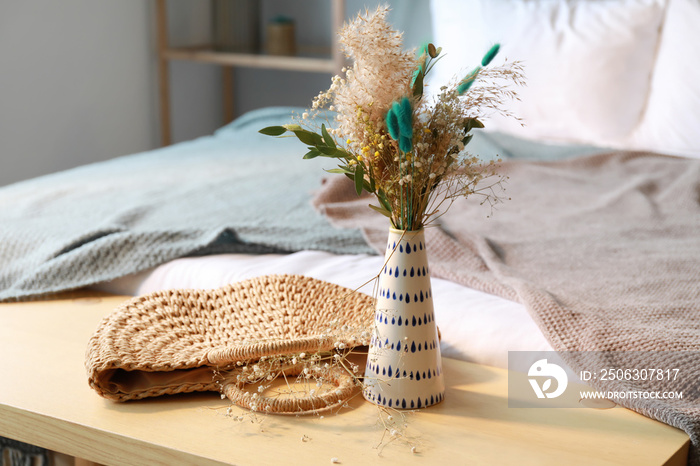 Vase with beautiful bouquet of dried flowers and wicker bag on wooden table in bedroom