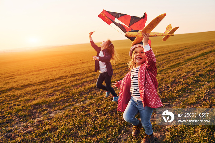 Two little girls friends have fun together with kite and toy plane on the field at sunny daytime