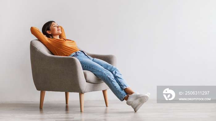 Young Asian woman sitting in comfy armchair, being lazy, feeling peaceful against white wall, panora