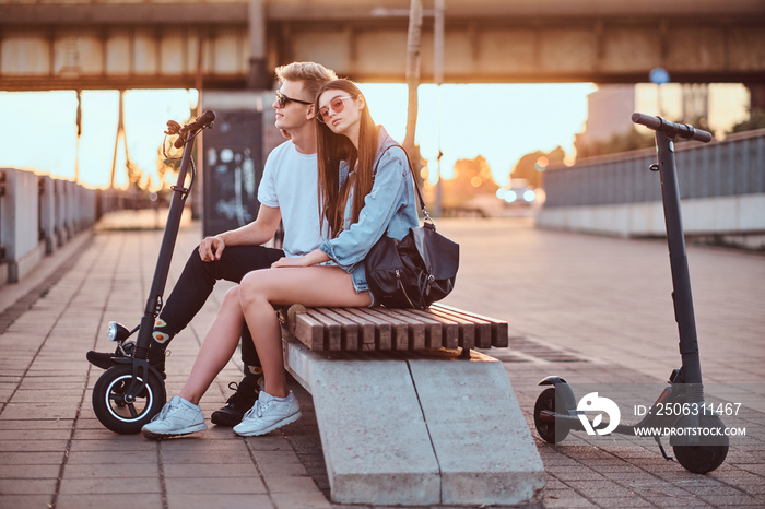 Young beautiful romantic couple with electric scooters is sitting on the bench near brige at sunset 