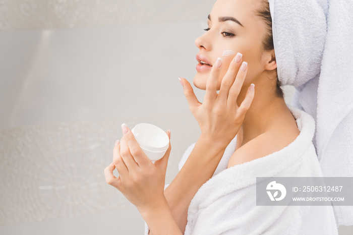 beautiful woman in bathrobe with towel on head applying face cream