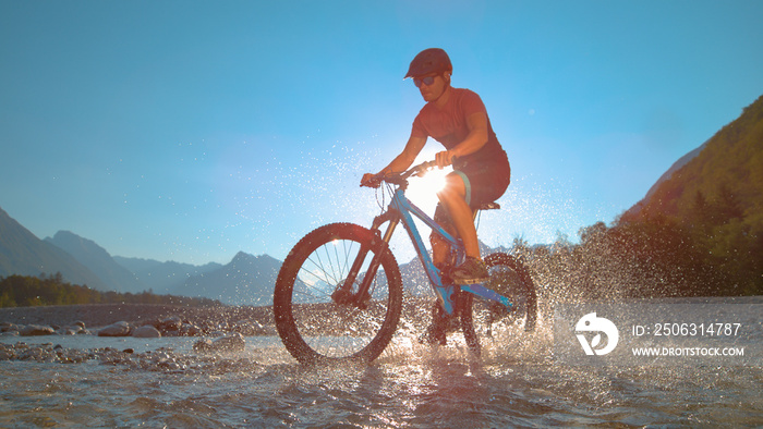 LOW ANGLE: Fit man rides an e-bike in the shallow river and splashes water.