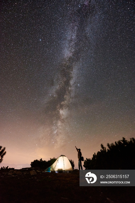 Silhouette of male traveler standing at night camp on the top of mountain beside illuminated tourist