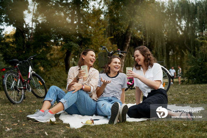 Three beautiful happy young women close friends relax in a green park on a blanket. Have fun laughin