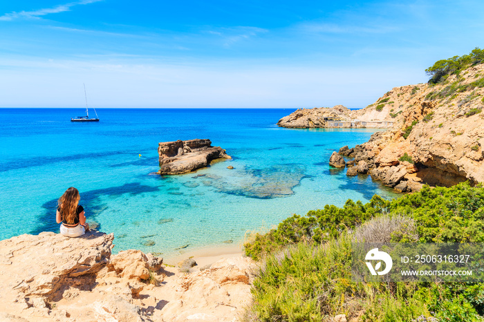 Unidentified young woman sitting on a rock and looking at beautiful Cala Tarida bay, Ibiza island, S