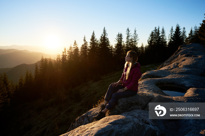 Young woman traveller sitting on big boulder on the top of mountain in the evening. Female tourist e