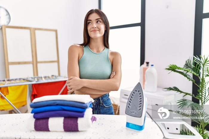 Young hispanic woman ironing clothes at laundry room smiling looking to the side and staring away th