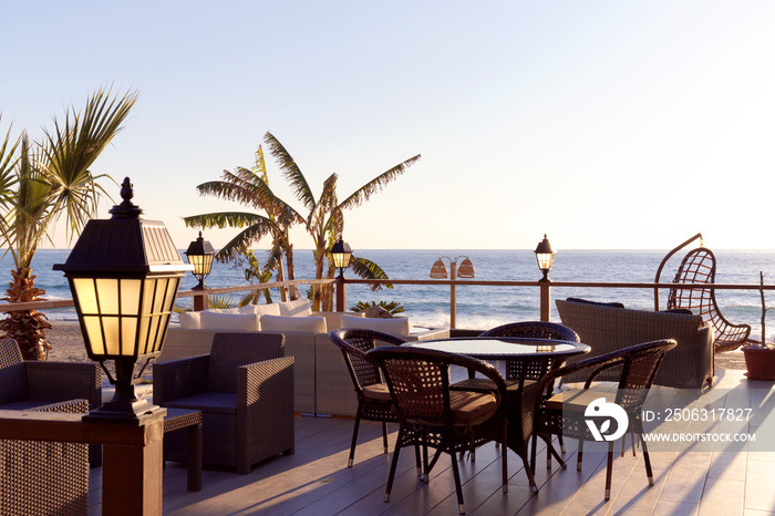 Cozy outdoor restaurant with lanterns in the rays of sun with sunset over sea on background, no peop