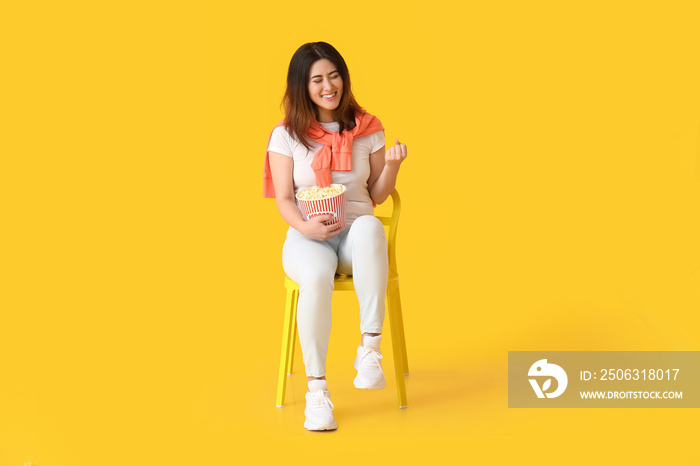 Happy young Asian woman with bucket of tasty popcorn sitting in chair on yellow background