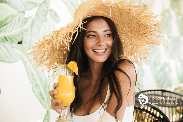 Photo of beautiful brunette woman in straw hat and summer clothes drinking orange juice in beach caf