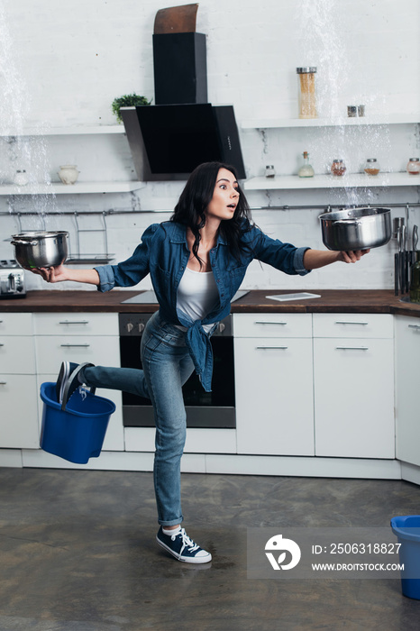 Frightened young woman with pots and buckets dealing with water leak in kitchen