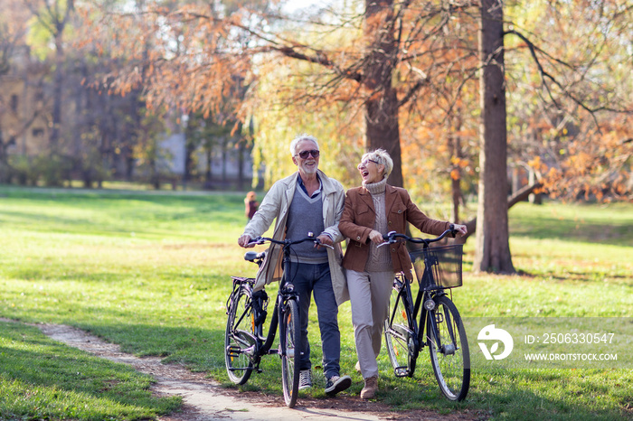 Mature fit couple pushing bicycles in public park talk and smile