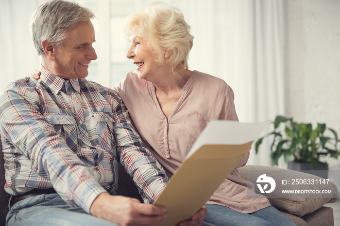 Mature husband and wife sitting in embrace with papers in hand. Their expressions are happy