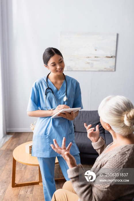 elderly woman gesturing while talking to smiling asian nurse holding digital tablet