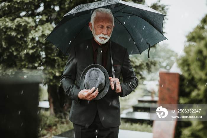 优雅悲伤的老人撑着伞站在雨中，在爱人的坟墓前悲伤
