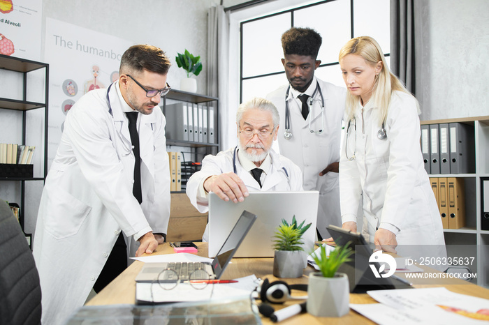 Multiracial therapists standing around aged professor that sitting at desk with modern laptop. Male 