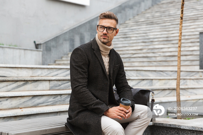 Image of handsome man 30s wearing eyeglasses, sitting on stairs with takeaway coffee while strolling