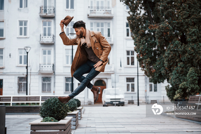 Holds hat and posing. Young male model in fashionable clothes is outdoors in the city at daytime