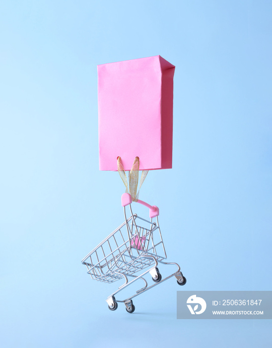 A pink shopping bag carries a shopping trolley on a blue background. Sales aesthetic shopping cart c