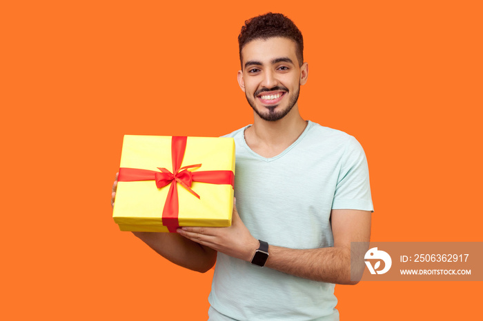 Portrait of positive young brunette man with beard in white t-shirt standing holding gift box and lo