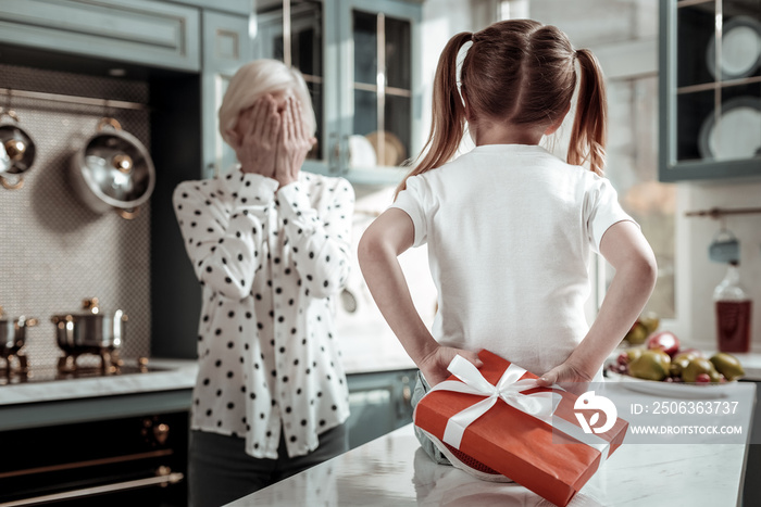 Little girl hiding big present and grandmother closing her eyes