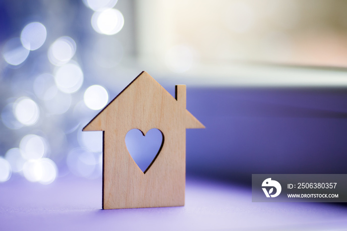 Wooden icon of house with hole in the form of heart on near window in daylight with bokeh lights on 