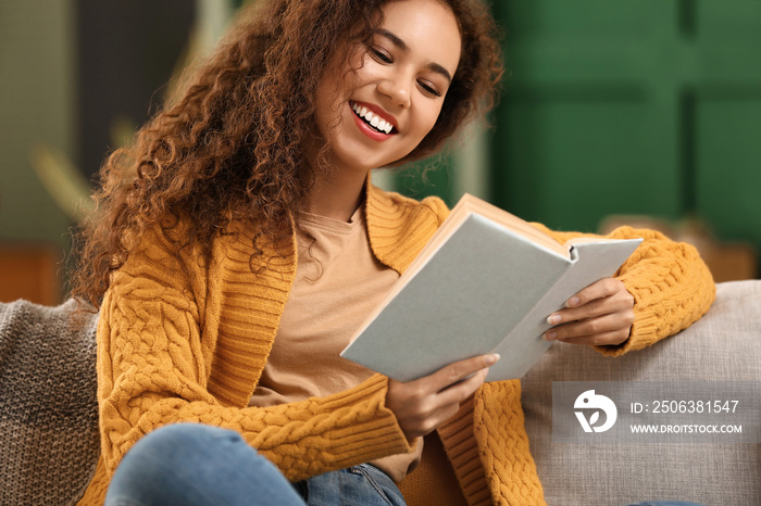 Young African-American woman in yellow cardigan reading book on sofa at home