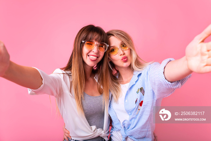 Photo of two young party women smiling and waving hand at camera while taking selfie photo on mobile