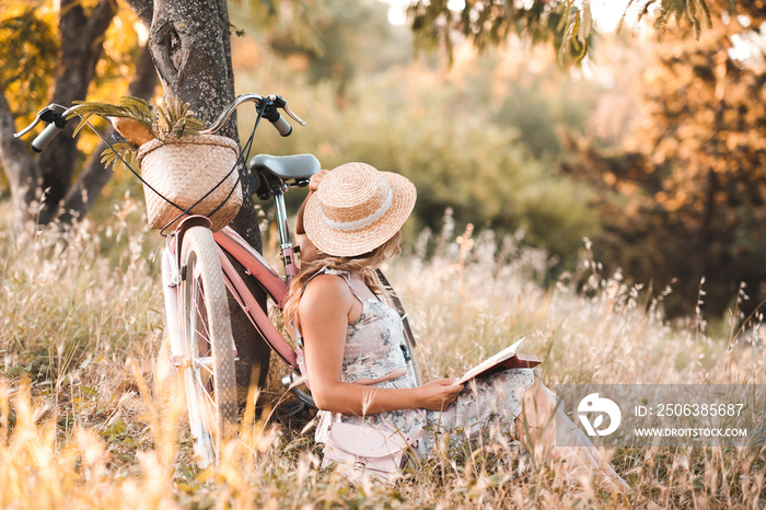 Woman sitting under tree in meadow with bike and book. Wearing straw hat and dress. Summer time.