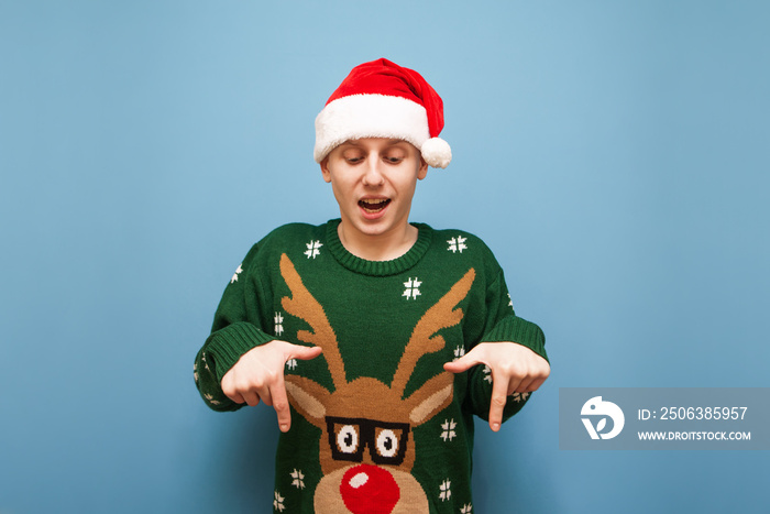 Cheerful guy in christmas clothes, looking down and showing spots to a blank. Young man in santa hat