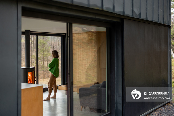 Young woman stands near fireplace at modern house on nature. View from outside through the window. C