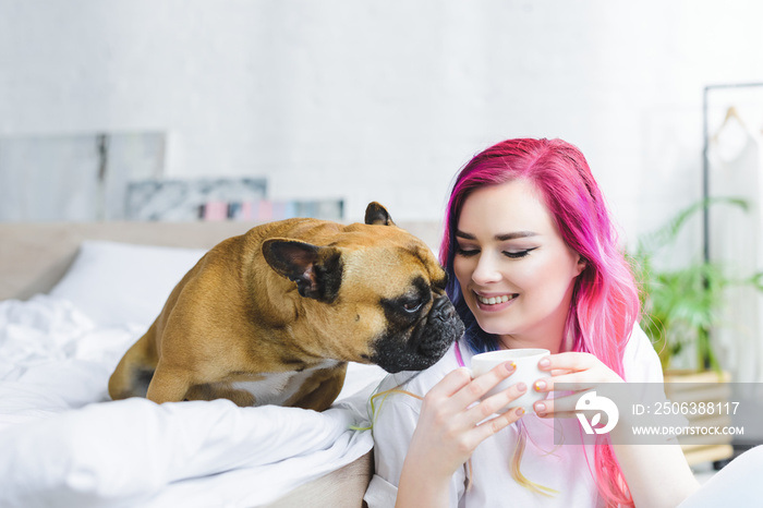 happy girl with colorful hair holding cup with coffee and cute bulldog sitting on bed and sniffing c