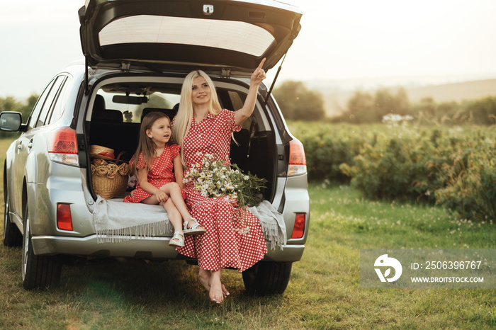 Young Mom with her Little Daughter Dressed Alike in Red Polka Dot Dress, Sitting in a Car Trunk in t