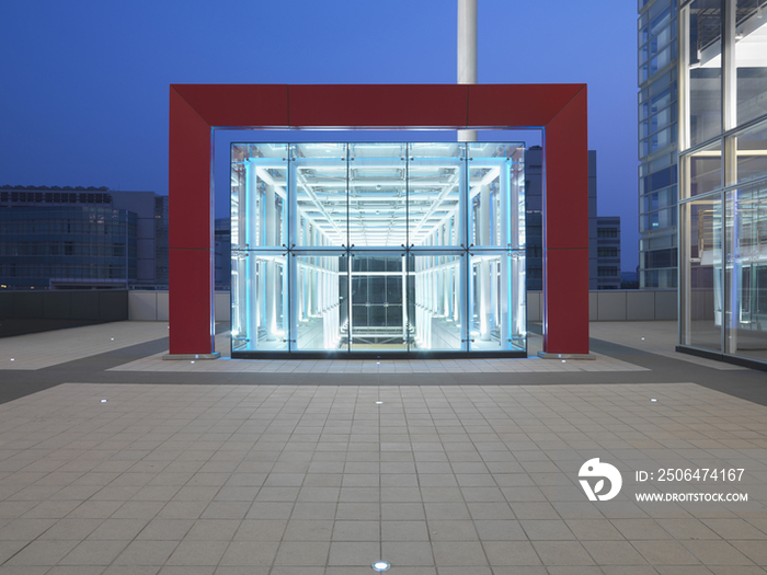 Red framed glass structure by office building against clear blue sky