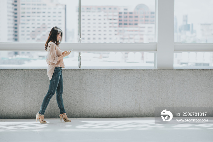 Beautiful Asian woman using smartphone for texting while walking in modern office building